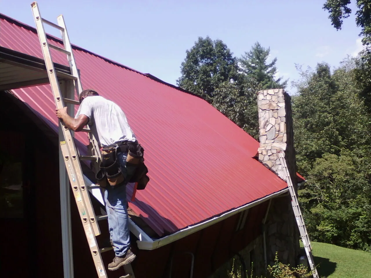 Roofer on a ladder beside a red metal roof and stone chimney — Soffit & Fascia Repair by Roofing Brothers in Chadds Ford